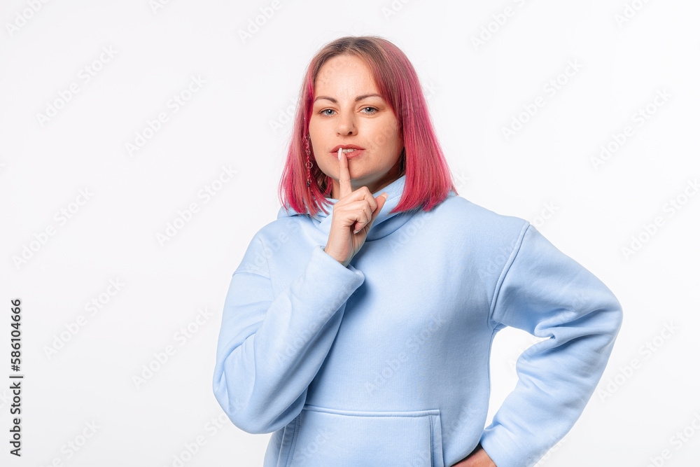 Serious woman holding finger at her lips asking not to make noise. Lovely female model with pink hair in a blue hoodie standing over white background Serious woman holding finger at her lips asking not to make noise. Lovely female model with pink hair in a blue hoodie standing over white background