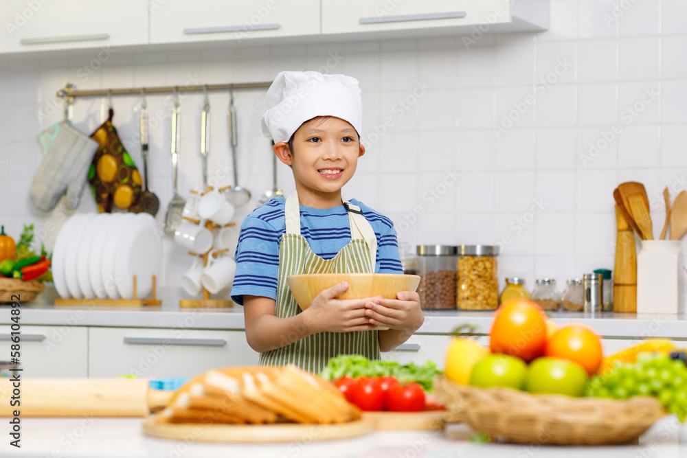 Millennial Asian young little boy chef wearing white tall cook hat and apron standing smiling holding wooden bowl posing taking photo while preparing cooking fresh meal at counter in home kitchen Millennial Asian young little boy chef wearing white tall cook hat and apron standing smiling holding wooden bowl posing taking photo while preparing cooking fresh meal at counter in home kitchen