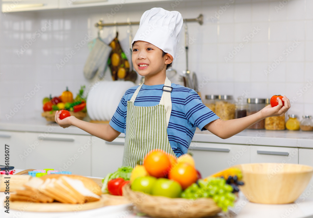 Millennial Asian little boy chef wearing tall white cook hat and apron standing holding red tomatoes posing in home kitchen full of fruits vegetables and sliced bread on cooking counter