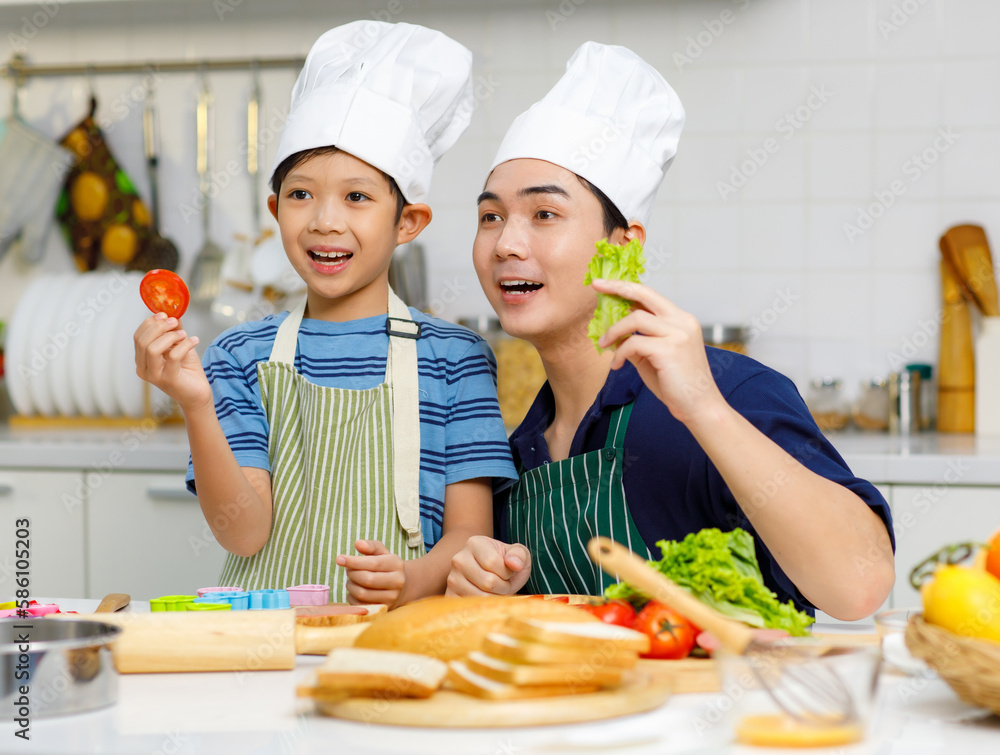 Millennial Asian little young boy chef wearing tall white cook hat and apron standing smiling holding sliced tomato posing together with father in home kitchen full of ingredients on cooking counter Millennial Asian little young boy chef wearing tall white cook hat and apron standing smiling holding sliced tomato posing together with father in home kitchen full of ingredients on cooking counter