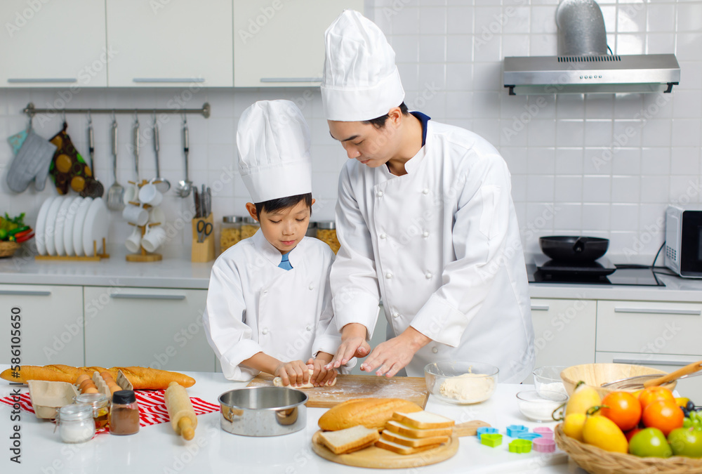 Asian young little boy chef in uniform with white tall cook hat standing learning preparing massaging flour dough on wooden cutting board while male cooking teacher helping teaching in home kitchen Asian young little boy chef in uniform with white tall cook hat standing learning preparing massaging flour dough on wooden cutting board while male cooking teacher helping teaching in home kitchen