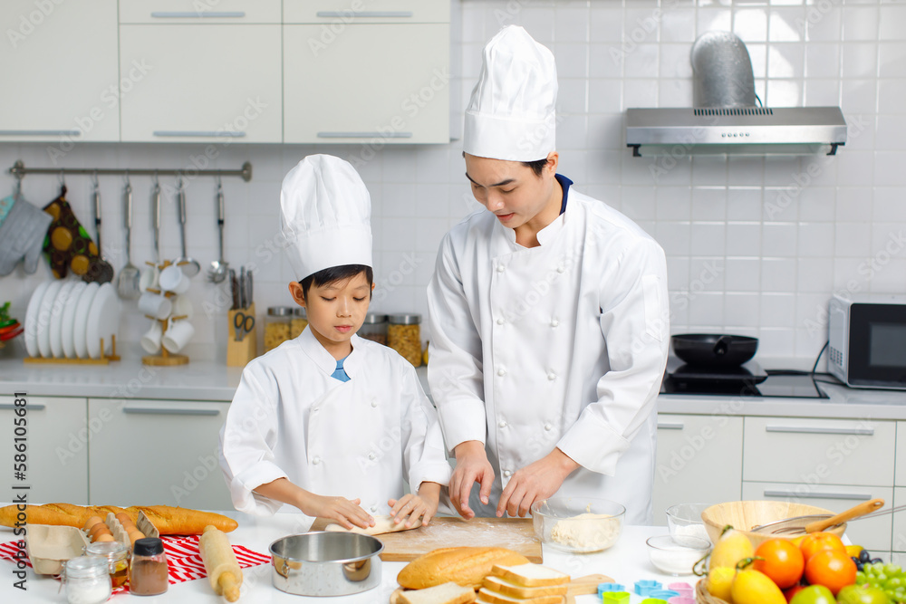 Asian young little boy chef in uniform with white tall cook hat standing learning preparing massaging flour dough on wooden cutting board while male cooking teacher helping teaching in home kitchen Asian young little boy chef in uniform with white tall cook hat standing learning preparing massaging flour dough on wooden cutting board while male cooking teacher helping teaching in home kitchen