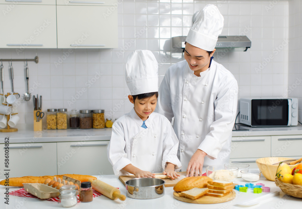 Asian young little boy chef in uniform with white tall cook hat standing learning preparing massaging flour dough on wooden cutting board while male cooking teacher helping teaching in home kitchen Asian young little boy chef in uniform with white tall cook hat standing learning preparing massaging flour dough on wooden cutting board while male cooking teacher helping teaching in home kitchen
