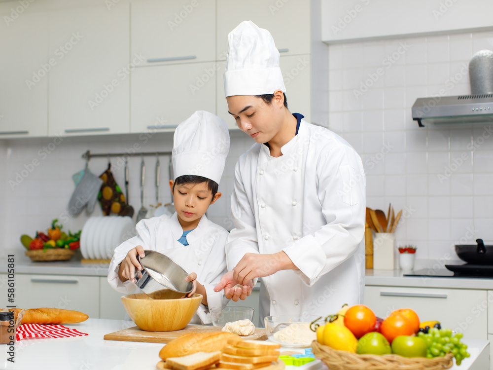 Asian young little boy pastry chef in white uniform with tall cook hat standing using stainless filter preparing sifting flour on wooden board while male cooking teacher helping teaching in kitchen Asian young little boy pastry chef in white uniform with tall cook hat standing using stainless filter preparing sifting flour on wooden board while male cooking teacher helping teaching in kitchen
