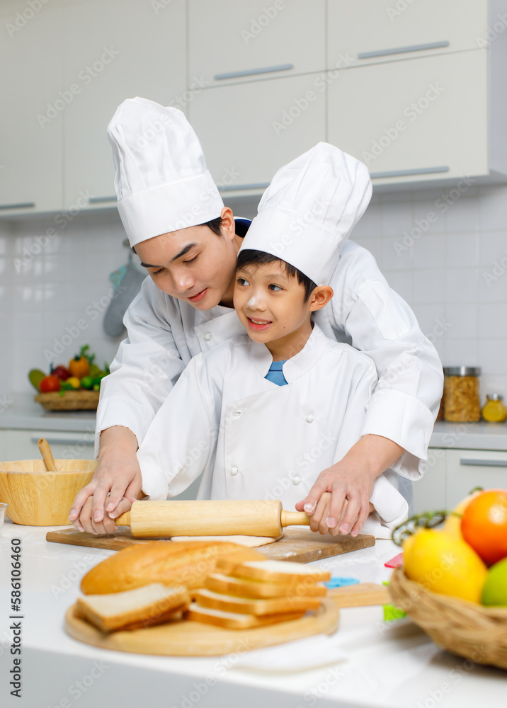 Asian young little boy chef in uniform with white tall cook hat standing learning preparing massaging flour dough on wooden cutting board while male cooking teacher helping teaching in home kitchen Asian young little boy chef in uniform with white tall cook hat standing learning preparing massaging flour dough on wooden cutting board while male cooking teacher helping teaching in home kitchen