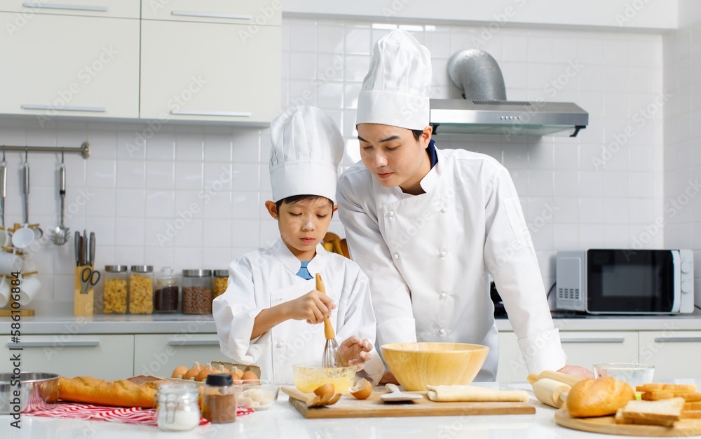 Asian young little boy pastry chef in white uniform with tall cook hat standing while male cooking teacher smiling help teaching in white kitchen. Asian young little boy pastry chef in white uniform with tall cook hat standing while male cooking teacher smiling help teaching in white kitchen.