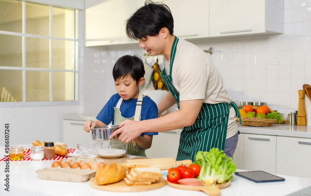 Asian young little boy chef wearing apron standing using stainless filter sifting white flour into glass bowl while father helping at counter full of baking equipment eggs breads tomatoes in kitchen
