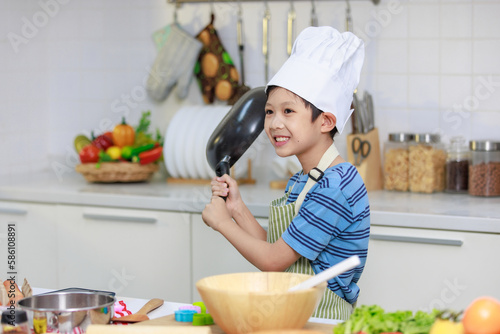 Millennial Asian young little boy chef wearing white tall cook hat and apron standing smiling holding cooking pan posing taking photo while preparing cooking fresh meal at counter in home kitchen