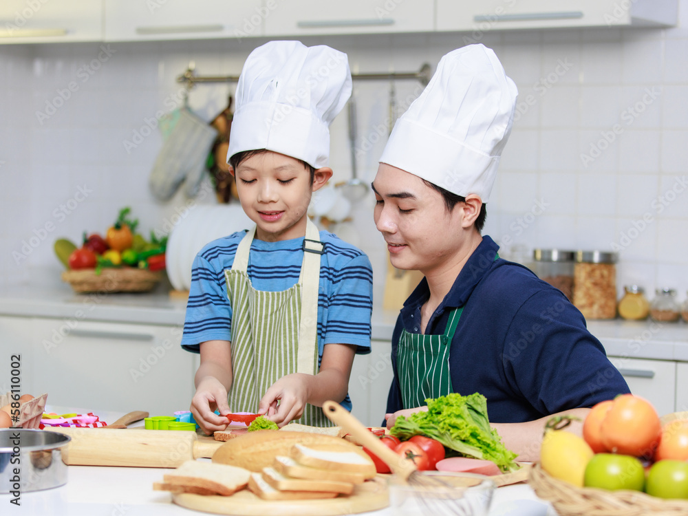 Asian young little boy chef wearing white tall cook hat and apron standing smiling holding putting sliced tomato on bread making preparing sandwiches while father helping teaching in home kitchen Asian young little boy chef wearing white tall cook hat and apron standing smiling holding putting sliced tomato on bread making preparing sandwiches while father helping teaching in home kitchen