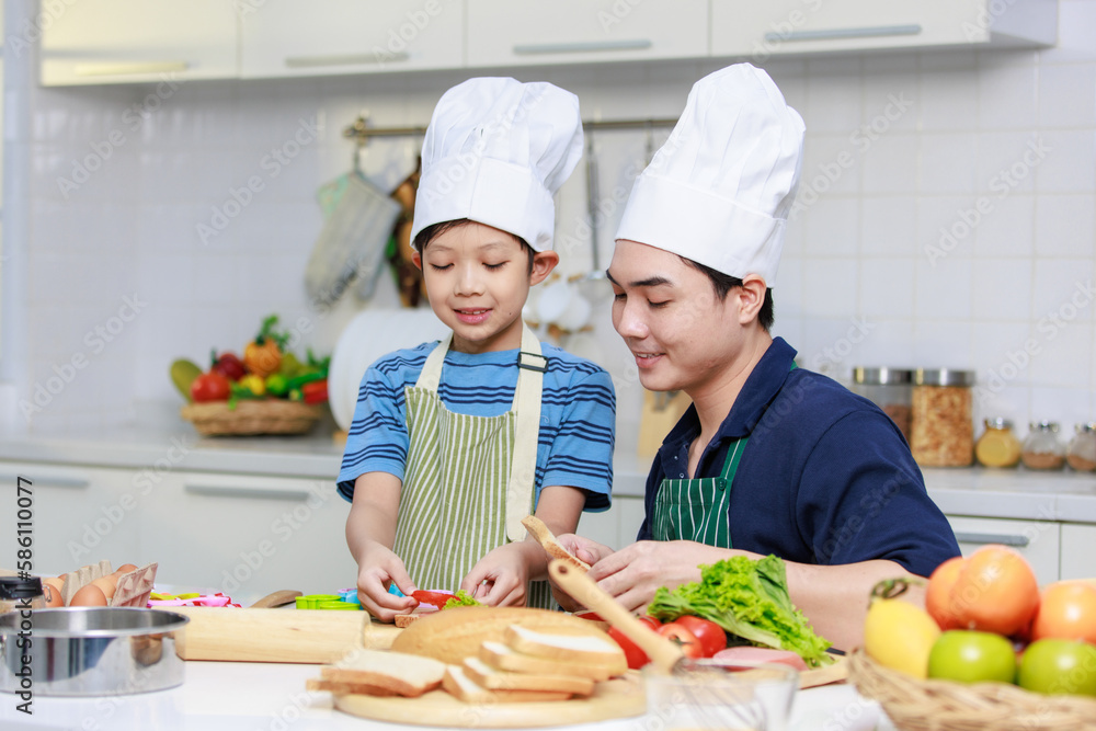 Asian young little boy chef wearing white tall cook hat and apron standing smiling holding putting sliced tomato on bread making preparing sandwiches while father helping teaching in home kitchen Asian young little boy chef wearing white tall cook hat and apron standing smiling holding putting sliced tomato on bread making preparing sandwiches while father helping teaching in home kitchen