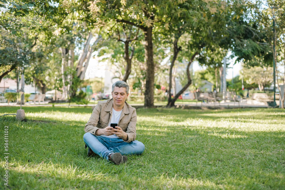 Gray-haired man looking at his cell phone sitting on the grass in a park. Gray-haired man looking at his cell phone sitting on the grass in a park.