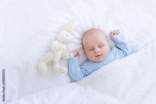 baby boy sleeping on the bed lying on his back with a stuffed toy hares in blue pajamas hands up, healthy newborn sleep