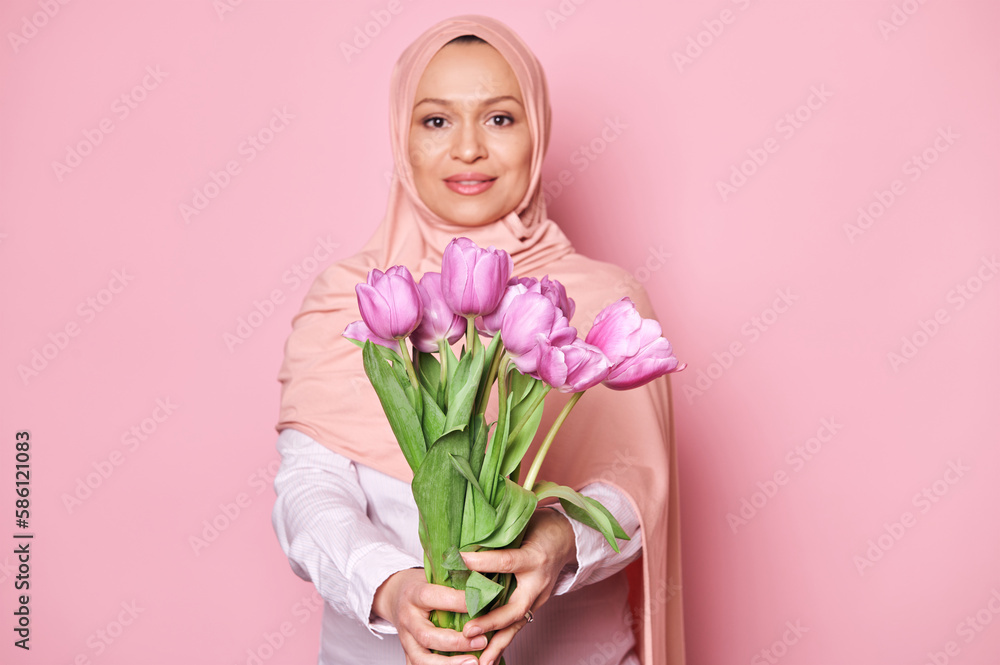 Details on a beautiful bouquet of pink and purple tulips in the hands of blurred Muslim woman, dressed in pink hijab, smiling looking at camera, isolated over color background. Spring holidays concept Details on a beautiful bouquet of pink and purple tulips in the hands of blurred Muslim woman, dressed in pink hijab, smiling looking at camera, isolated over color background. Spring holidays concept