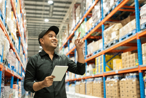 Warehouse worker wearing a hat and black shirt hands holding tablet check stock on tall shelves in warehouse storage. Asian auditor or staff work looking up stocktaking inventory in warehouse store.