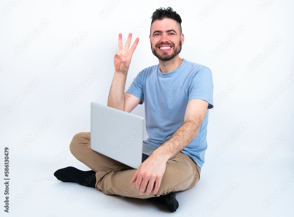 Young caucasian man with laptop sitting over white studio showing and pointing up with fingers number three while smiling confident and happy. Young caucasian man with laptop sitting over white studio showing and pointing up with fingers number three while smiling confident and happy.