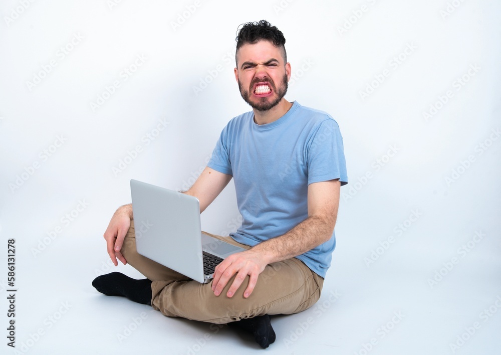 Young caucasian man with laptop sitting over white studio keeps teeth clenched, frowns face in dissatisfaction, irritated because of much duties. Young caucasian man with laptop sitting over white studio keeps teeth clenched, frowns face in dissatisfaction, irritated because of much duties.