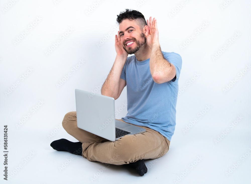 Young caucasian man with laptop sitting over white studio Trying to hear both hands on ear gesture, curious for gossip. Hearing problem, deaf Young caucasian man with laptop sitting over white studio Trying to hear both hands on ear gesture, curious for gossip. Hearing problem, deaf