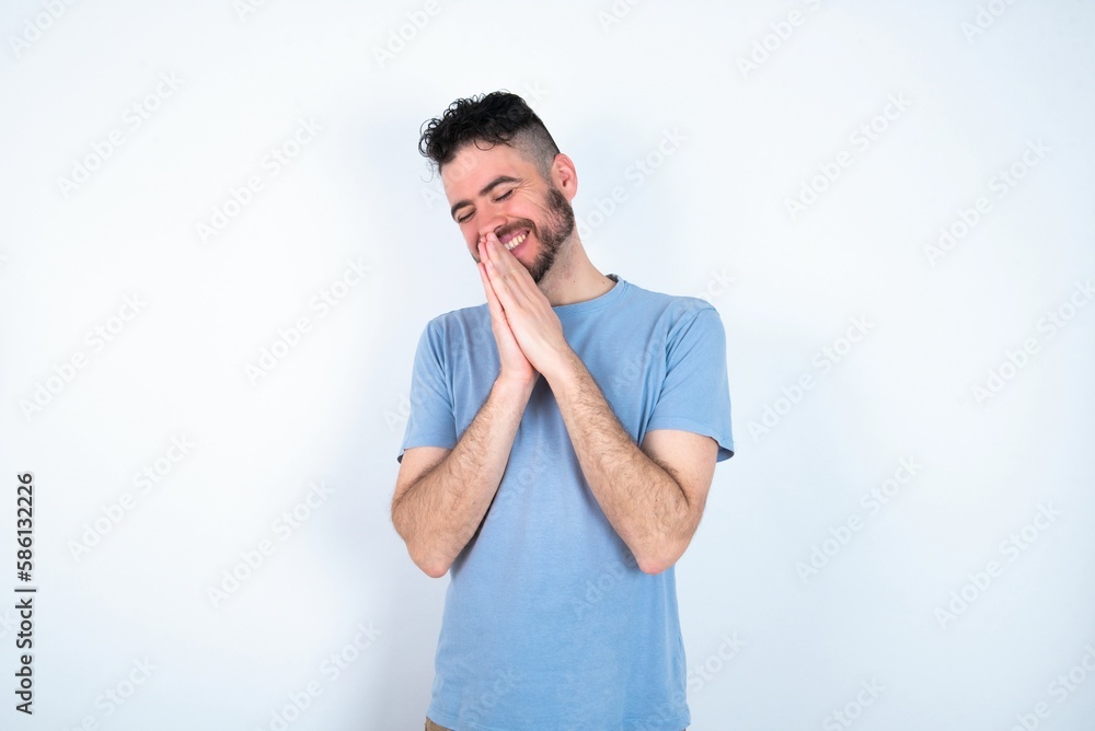 Overjoyed Young caucasian man wearing blue T-shirt over white background laughs joyfully and keeps palms pressed together hears something funny Overjoyed Young caucasian man wearing blue T-shirt over white background laughs joyfully and keeps palms pressed together hears something funny