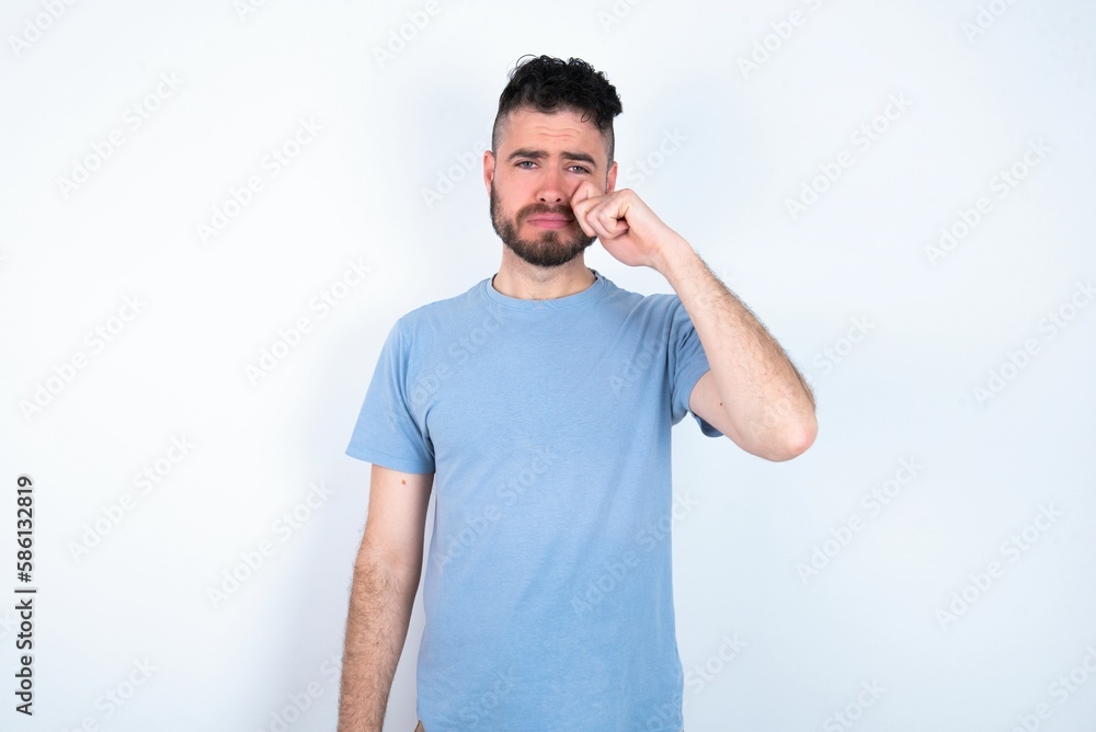 Disappointed dejected Young caucasian man wearing blue T-shirt over white background wipes tears stands stressed with gloomy expression. Negative emotion Disappointed dejected Young caucasian man wearing blue T-shirt over white background wipes tears stands stressed with gloomy expression. Negative emotion