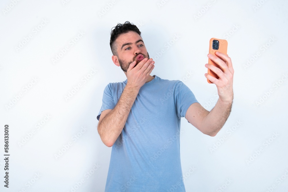 Young caucasian man wearing blue T-shirt over white background blows air kiss at camera of smartphone and takes selfie, sends mwah via online call. Young caucasian man wearing blue T-shirt over white background blows air kiss at camera of smartphone and takes selfie, sends mwah via online call.