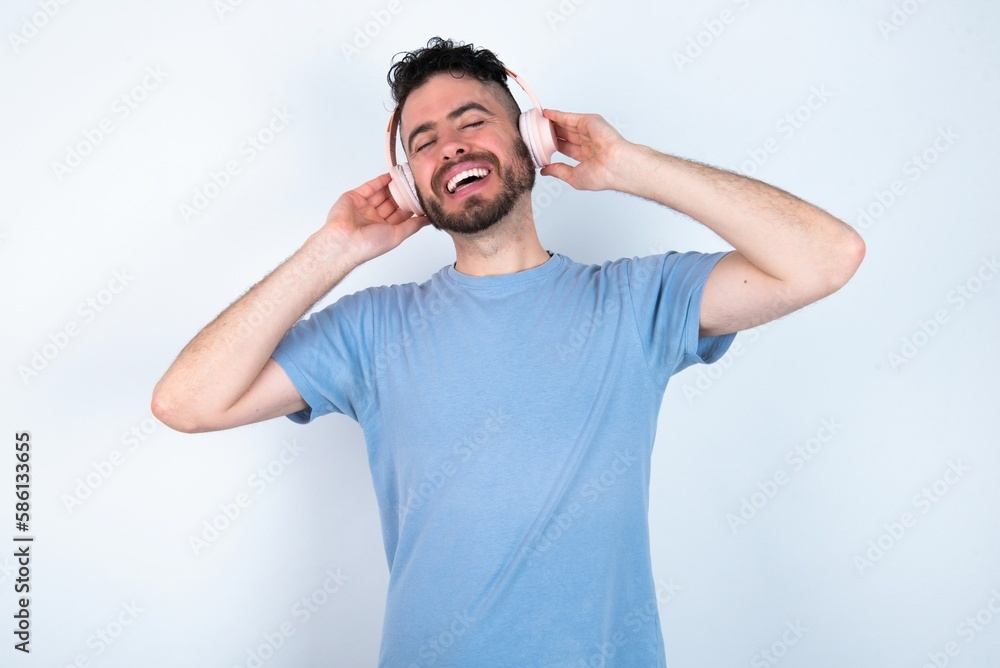 Young caucasian man wearing blue T-shirt over white background smiles broadly feels very glad listens favourite music track via wireless headphones closes eyes. Young caucasian man wearing blue T-shirt over white background smiles broadly feels very glad listens favourite music track via wireless headphones closes eyes.