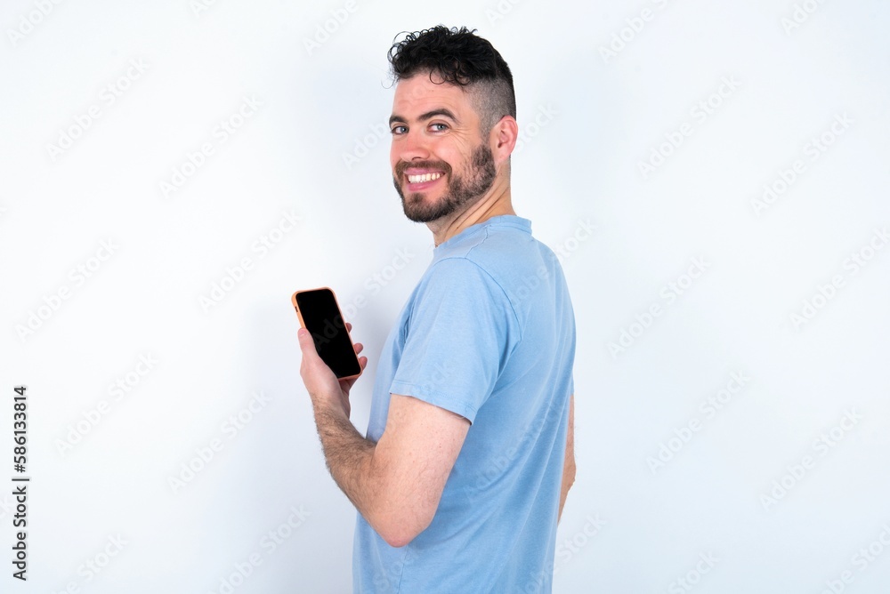 Rear view photo portrait of Young caucasian man wearing blue T-shirt over white background using smartphone smiling Rear view photo portrait of Young caucasian man wearing blue T-shirt over white background using smartphone smiling