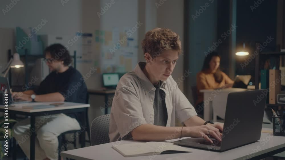 Young short haired businesswoman in casualwear using laptop and making notes at desk in the office, overworking during night, colleagues in the background Young short haired businesswoman in casualwear using laptop and making notes at desk in the office, overworking during night, colleagues in the background