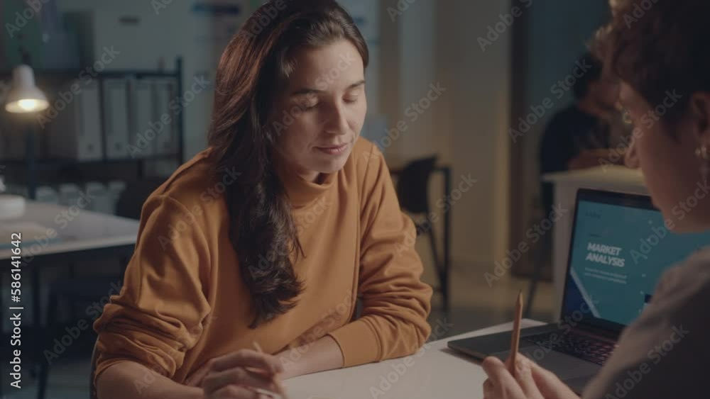 Young businesswoman discussing project with female colleague in the office, working after hours during the night. Overworking concept, over the shoulder tilt up shot Young businesswoman discussing project with female colleague in the office, working after hours during the night. Overworking concept, over the shoulder tilt up shot