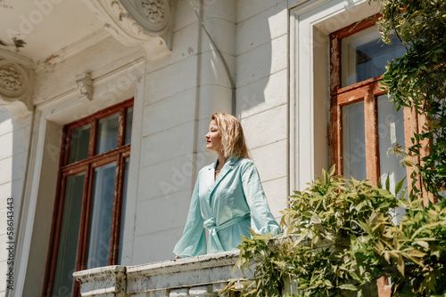 A woman on a balcony in a long blue dress stands near a low granite stone fence against the background of an old stone house