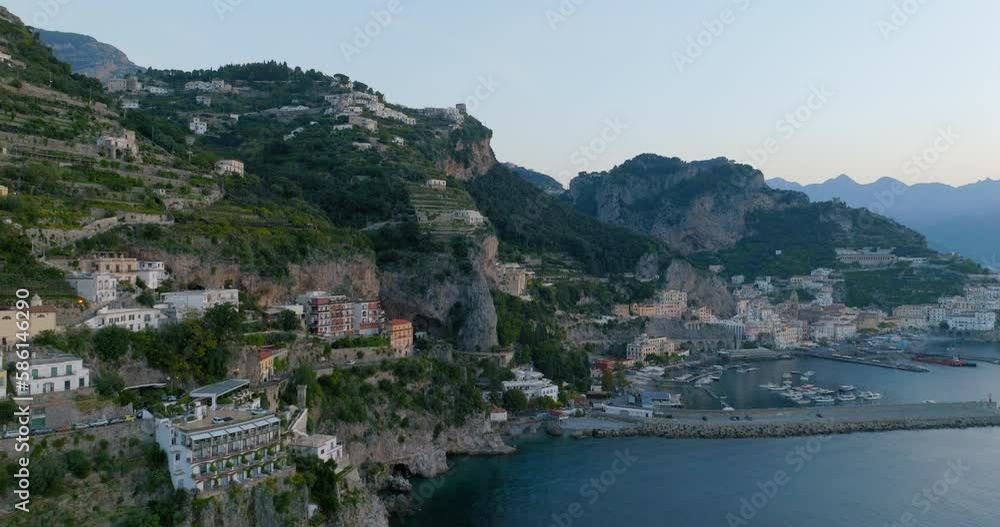 Aerial Forward Shot Of Hotel Buildings On Rock Formations By Nautical Vessels Moored At Harbor During Sunset - Amalfi, Italy Aerial Forward Shot Of Hotel Buildings On Rock Formations By Nautical Vessels Moored At Harbor During Sunset - Amalfi, Italy