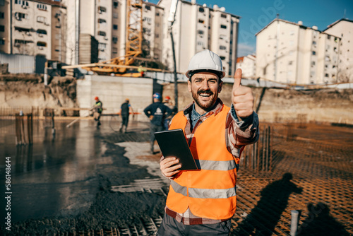 A constructor is standing on building foundation with tablet in his hands and giving thumbs up and smiling at the camera. The workers in background are concreting foundation. © dusanpetkovic1