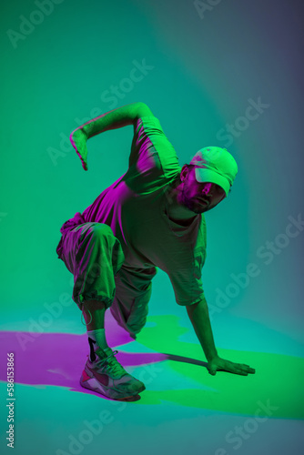 Cool trendy professional hip hop dancer man with cap in fashion clothes dancing in a colorful studio with green and pink lights