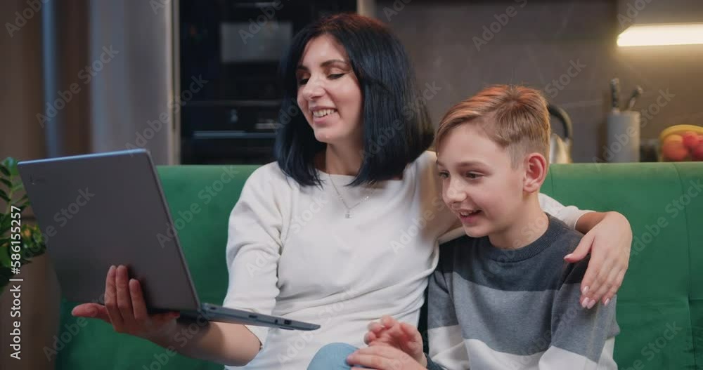 Beautiful woman with cute 9-year-old boy sitting together on couch holding laptop while talking on video chat with family. Happy family and modern technology Beautiful woman with cute 9-year-old boy sitting together on couch holding laptop while talking on video chat with family. Happy family and modern technology