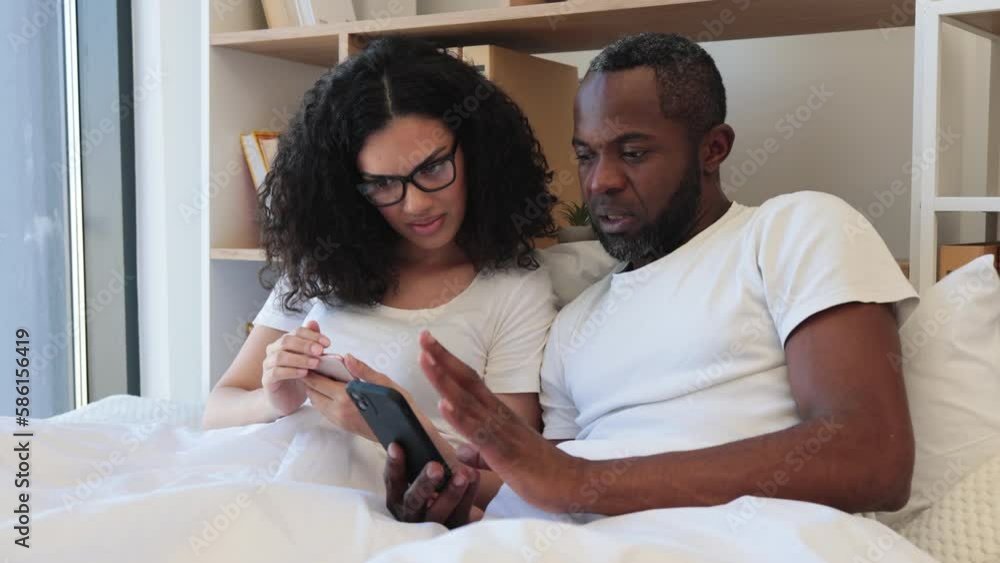 Serene multiracial family of two using mobile phones while lazing in soft bed during daytime at home. Confident adult man and bespectacled woman reading online article paying attention to details.