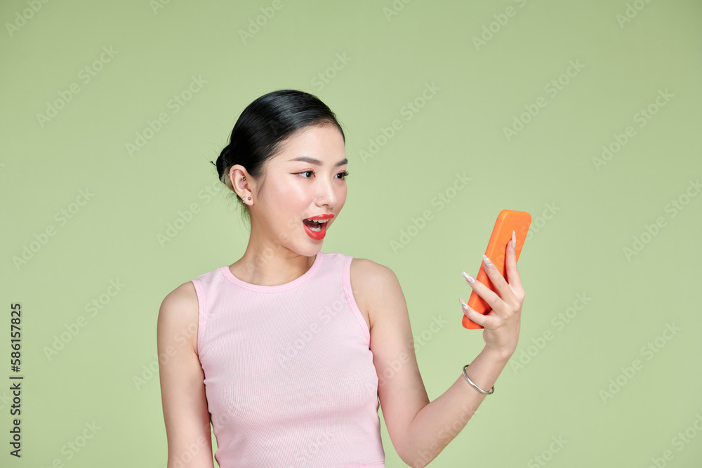 Young beautiful Asian woman feeling happy or surprise shock and holding smartphone over green background Young beautiful Asian woman feeling happy or surprise shock and holding smartphone over green background