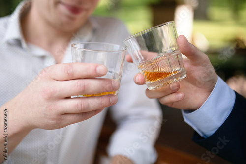 Two men clanging glasses of whiskey or bourbon. Hands of men holding glasses with alcohol. Friendly outdoor party. Glass clinking at wedding. Cheers
