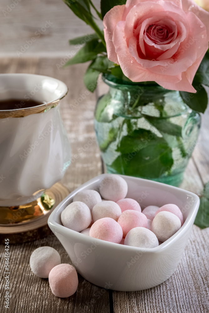 Round white and pink candies in a vase, a rose in a glass vase and a cup of tea on a wooden table. Postcard. Photo