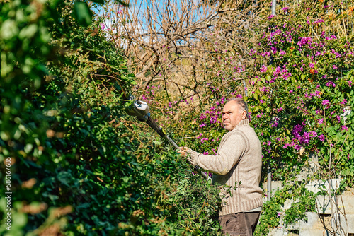 Mature man cutting shrub with hedge trimmer. Male gardener working with professional garden equipment in backyard, using modern electric trimmer for work outdoors.