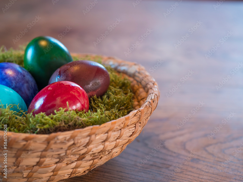 Fototapeta premium Various coloured decorated easter eggs in wicker basket nest with moss and colored Easter eggs on beige wooden bokeh background. For Happy Easter holiday card. Selective focus, copy space