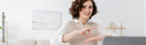 happy teacher with curly hair showing sign language gesture during online lesson on laptop, banner. photo