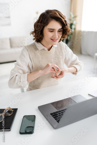 happy young teacher showing interpretation sign on body language near laptop and smartphone at home.