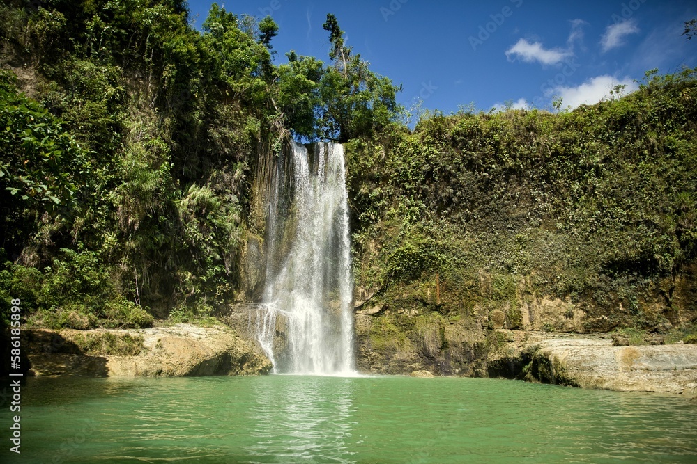 Fototapeta premium The idyllic Camugao Waterfall in Siquijor in the Philippines that flow into a natural pool of water surrounded by the light-lit rainforest.