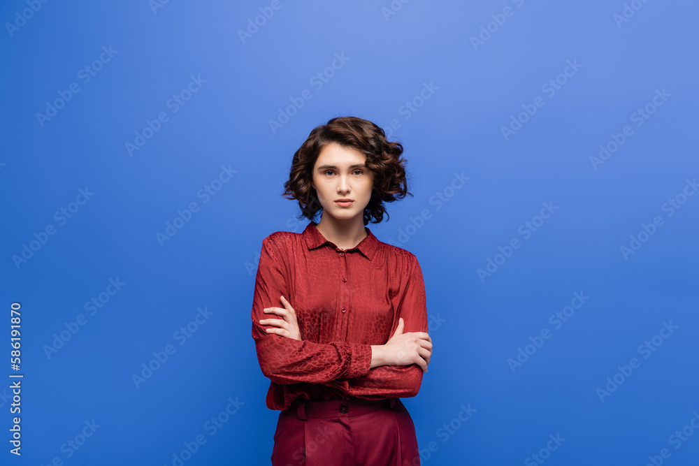 pretty language teacher with wavy hair looking at camera while standing with crossed arms isolated on blue. pretty language teacher with wavy hair looking at camera while standing with crossed arms isolated on blue.