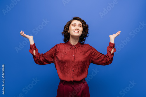 Obraz na płótnie smiling young woman with short curly hair gesturing while looking away isolated on blue