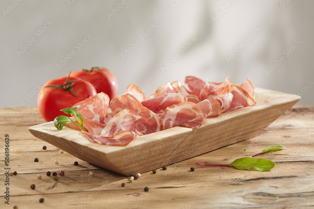 meat delicacy with green leaves and tomatoes on a wooden background. dry-cured pork on a wooden table. meat with a layer of fat under sunlight. traditional snack in rustic style. meat delicacy with green leaves and tomatoes on a wooden background. dry-cured pork on a wooden table. meat with a layer of fat under sunlight. traditional snack in rustic style.
