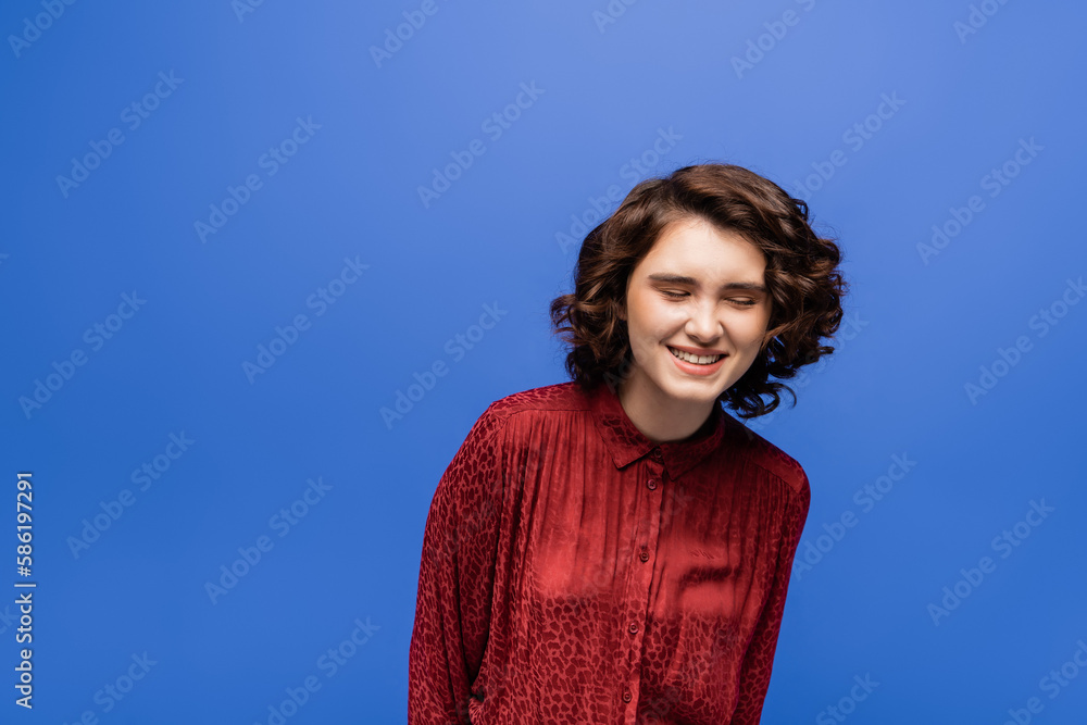 cheerful language teacher in red blouse with animal print smiling while closing eyes isolated on blue. cheerful language teacher in red blouse with animal print smiling while closing eyes isolated on blue.