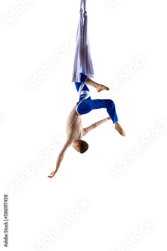 Artistic male acrobat, professional aerial gymnast hanging upside down on aerial silk against white studio background. Beautiful tricks. Concept of art, sportive lifestyle, hobby, action and motion