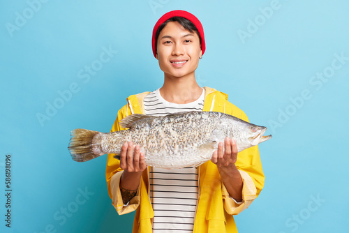 Young Asian fisherman standing and holding fish with happy expression emotions dressed in red hat and yellow fishing jacket isolated over blue background. People emotions concept