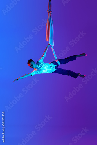 Acrobat, professional male aerial gymnast hanging upside down on aerial silk against gradient blue purple background in neon light. Concept of art, sportive lifestyle, hobby, action and motion, beauty