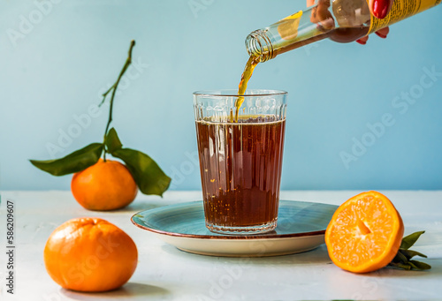 Chinotto refreshing citrus drink in a glass, woman's hand holding bottle and pouring drink to the glass, light surface with blue background